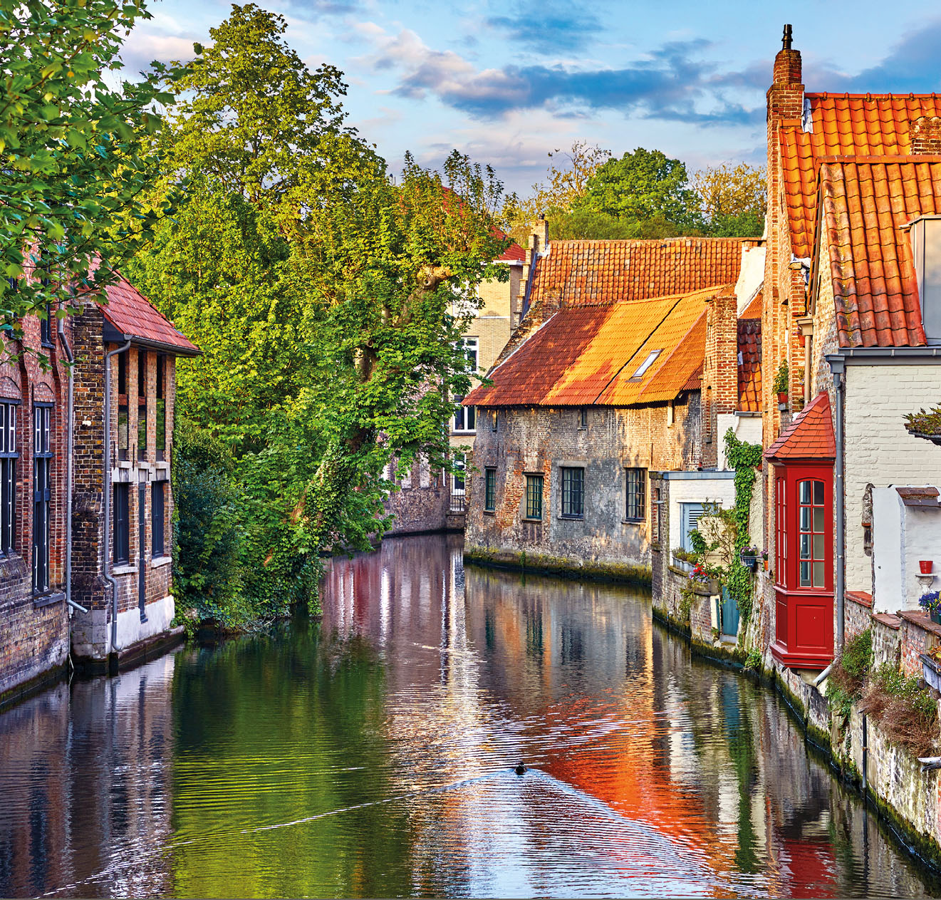 Bruges, Belgium. Medieval ancient houses made of old bricks at water channel with boats in old town. Summer sunset with sunshine and green trees. Picturesque landscape.