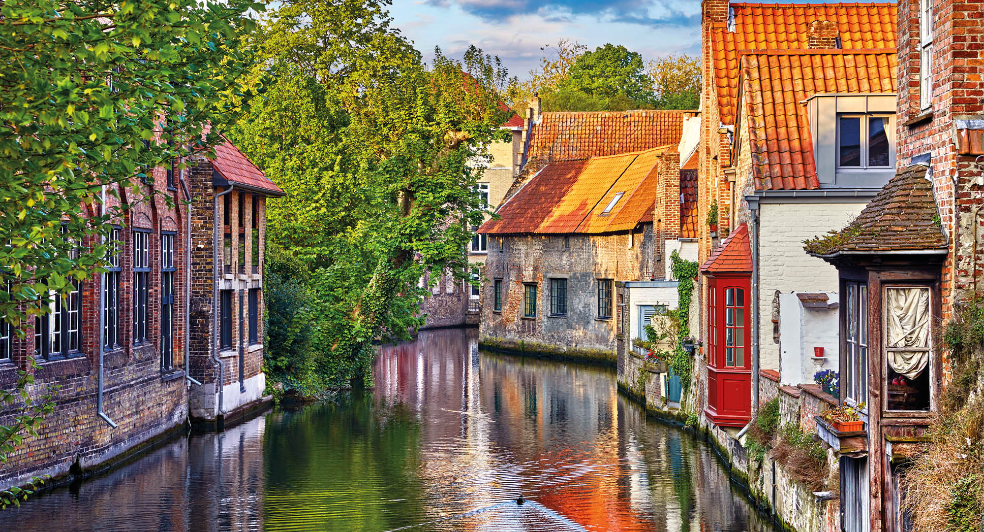 Bruges, Belgium. Medieval ancient houses made of old bricks at water channel with boats in old town. Summer sunset with sunshine and green trees. Picturesque landscape.