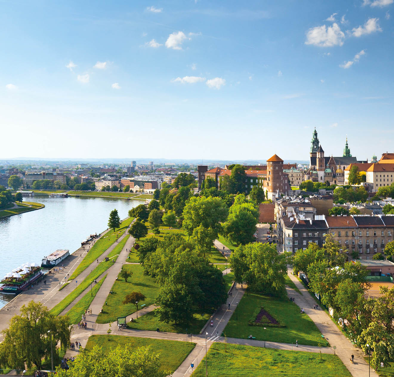 Daytime panorama of Krakow, Poland, featuring Wawel Castle. The Vistula River flows along the left side of the image, and the castle is visible on the right, near the horizon.  There is a blue sky with sparse, wispy clouds above the horizon and a manicured lawn intersected by walking paths in the foreground.