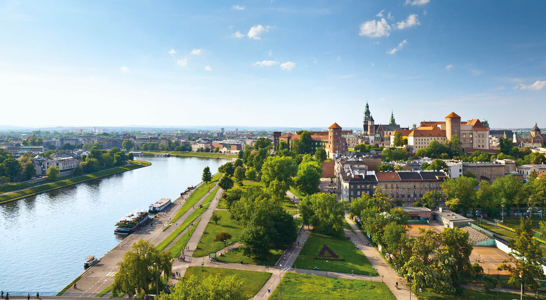Daytime panorama of Krakow, Poland, featuring Wawel Castle. The Vistula River flows along the left side of the image, and the castle is visible on the right, near the horizon.  There is a blue sky with sparse, wispy clouds above the horizon and a manicured lawn intersected by walking paths in the foreground.